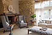 Reading chair in front of open fireplace in interior with enormous window and rustic stone walls
