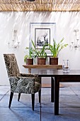 Gepolsterter Sessel im Retro-Look vor Holztisch gegenüber Wand mit Bild zwischen Kerzenhaltern auf überdachter Terrasse