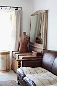 Traditional, brown leather sofa next to wooden trunk and tailor's dummy in front of framed mirror in corner of room next to window