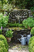 Zinc watering cans in front of stone-walled fountain with stone sheep's head as water spout