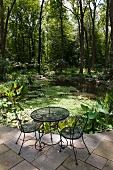 Wire mesh garden table and chairs on stone-flagged terrace with view of pond in woodland garden