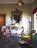 Überladener Konsolentisch und Wandspiegel, Polstersessel mit Goldfüssen und bunt gemusterter Stoff mit Totenkopf auf einem Stuhl