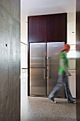 Offener Grundriss in puristischem Wohnhaus mit Sichtbetonwänden - Person läuft vor Einbaukühlschrank mit Edelstahlfronten