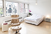 Minimalistisches Schlafzimmer mit hellem Sessel-Klassiker und passendem Fussschemel vor Doppelbett am Panoramafenster