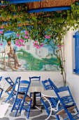 Chairs and tables in corner of terrace in front of mural with bucolic motif below climber-covered pergola