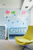 Wolken und Gleitschirmflieger auf himmelblauer Wand hinter Babybett; lindgrüner Drehsessel im Vordergrund