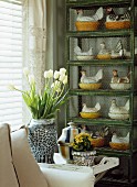 Bouquet of white tulips in ceramic vase on tray in front of china hen figurines on cage-like shelving