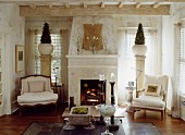 Open fireplace flanked by armchairs in front of decorative bushes in vases on columns against windows in country-style interior