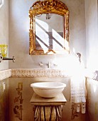 Antique stone basin on washstand below gilt-framed mirror in niche in bathroom with pattern of sunlight and shadow on wall
