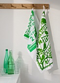 Printed, green and white dish cloths on wall hooks above a kitchen counter with bottles