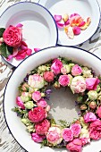 Kranz mit rosaroten Rosen in einer Emailleschüssel und Blütenblätter auf Tellern