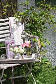 Tablett mit zarten Rosensträusschen in Wassergläsern und Einmachglas mit Schleife auf altem Gartenstuhl
