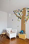 Kinderzimmer mit Babybett vor einem auf die Wand aufgemalten Baumstamm mit stilisierten Blättern