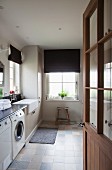 View into utility room with washing machine, dark Roman blinds and tiles in pale, natural shades