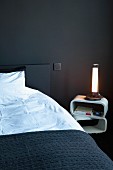Bed with black bedspread and lit table lamp on retro bedside table in bedroom with black-painted wall