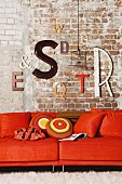 Decorative letters on brick wall and pendant lamp with cage-like lampshade above orange sofa with 70s scatter cushions
