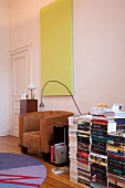 Mehrere Bücherstapel auf Boden neben hellbraunem Ledersessel an Wand vor hellgelbem monochromem Bild