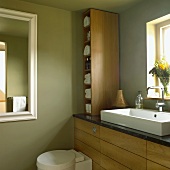 Bathroom with green walls, wooden furniture, sink in front of the window, toilet and wall mirror