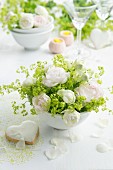 Arrangement of white roses and lady's mantle in china bowl next to heart-shaped, iced biscuit