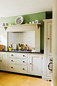 Kitchen counter in country-house kitchen with white base units and integrated gas hob below extractor hood on green wall