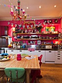 Kitchen counter along hot pink wall with long wooden shelves; simple wooden table with colourful retro chairs in foreground
