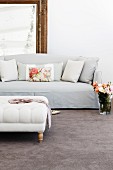 Glass vase of roses on grey velour carpet next to elegant, pale sofa with scatter cushions and ottoman with wooden legs in foreground