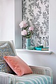 Pastellfarbene Kissen auf Sessel mit weißem Gestell und Geflecht neben Wand mit Blumenstrauss in tapezierter Nische