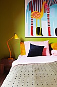 Tagesdecke und Kissen auf schlichtem Bett und Retro-Tischleuchte auf Nachttisch vor gelbgrüner Wand mit Bild