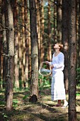 Young woman in romantic, white summer clothing with wicker basket in pine forest