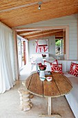 Rustikaler Holztisch und weisses Sofa auf überdachter Terrasse mit weissen, bodenlangen Vorhängen; Blick durch offene Tür ins Esszimmer