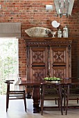Dark wooden dining table and chairs in front of antique, carved cabinet and exposed brick wall