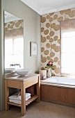 Simple washstand with white basin below mirror and bathtub against wall with retro wallpaper