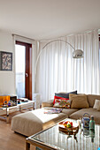 Helles Wohnzimmer mit beigem Sofa, Bogenleuchte, Glas-Couchtisch und weißen Gardinen