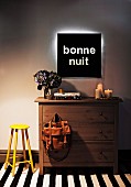 Hand-crafted black wall lamp with backlit lettering on white wall above wooden chest of drawers on black and white striped rug