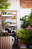 Woman reading on loft balcony with herbs on shelves, trellis and flowering plants in wooden planters