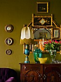 Table lamps, various glass vessels and vase of roses on chest of drawers in front of arrangement of mirrors on green-painted wall