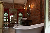 Mirror with Indian wooden frame, draped saris, free-standing bathtub and antique washstand in shabby-chic bathroom with rustic brick walls