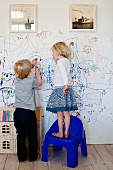 Two children scribbling on a white wall below framed photographs