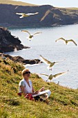 Frau im Schneidersitz auf Wiese über Meeresklippe beim Zeichnen fliegender Möwen
