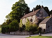Traditionelles, englisches Landhaus mit Natursteinfassade; blühende Fassadenberankung und dichte Gehölze hinter Steinmauer