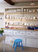 Blauer Holzschemel vor rustikaler Küchenzeile mit Unterschrank und weisses Geschirr im Wandregal