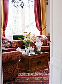 Blick durch offene Tür auf Holztruhe mit Blumenstrauss und Sesseln vor Fenster im traditionellen Wohnzimmer