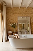Free-standing bathtub with wooden shelf and mirror leaning against stone wall in ensuite bathroom