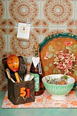 Ethnic wooden spoon in old tin can, bottle of home-made vinegar with decorative tag and drinking bowl on 70's board in front of tray with rustic, floral painted motif; 70's tiles in background