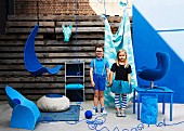 Boy and girl in blue clothing standing amongst furniture for child's bedroom in shades of blue in front of rustic wooden wall