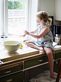 Kleines Mädchen sitzt auf Küchenschrank neben Backzutaten beim Fenster