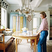 Woman setting table in dining area with Biedermeier furniture and painted farmhouse cupboard in background