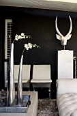 White flowers in metal vases on tray and white chairs next to animal-head sculpture against black wall