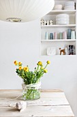 Bouquet of ranunculus in glass vase with decorative cord on wooden table; detail of pendant lamp in foreground and shelving in niche in background
