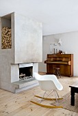 Eames rocking chair in front of concrete chimney breast with open fireplace and niche for firewood; piano with swivel stool in background